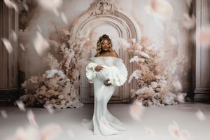 Dreamy maternity photo with white gown and floral arch backdrop in soft lighting.