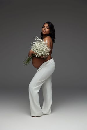 Modern maternity portrait with white pants and baby's breath bouquet against grey backdrop.