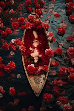 Dramatic photo of figure lying in wooden boat surrounded by floating red roses on dark water.