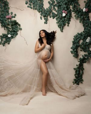 Maternity portrait with sheer fabric against wall adorned with green foliage.