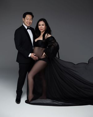 Glamorous maternity portrait with flowing black gown and tuxedo against grey backdrop.