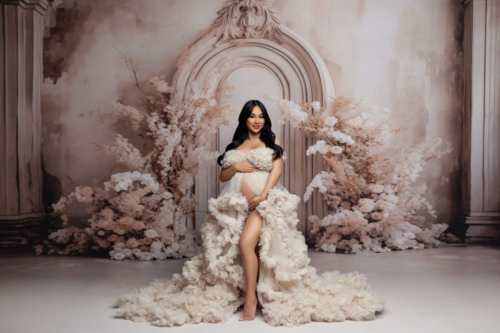 Romantic maternity portrait with cream ruffled gown amid floral arch and dried pampas grass.
