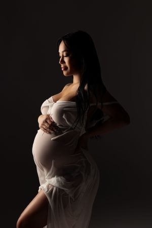 Dramatic side-lit maternity silhouette in white flowing fabric against dark background.