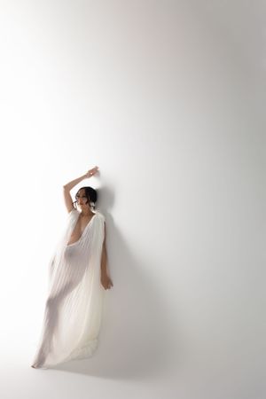 Ethereal figure in flowing white dress poses gracefully against light background.