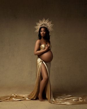 Artistic maternity photo featuring gold fabric and crown-like headpiece in dramatic studio lighting.