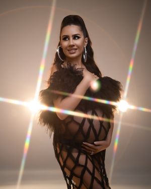 Fashion model wearing black mesh gown with feather details and statement earrings against glowing backdrop.