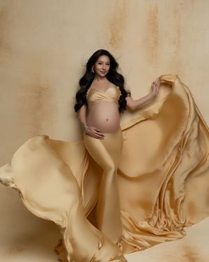 Elegant maternity portrait with billowing golden silk fabric against warm beige backdrop.