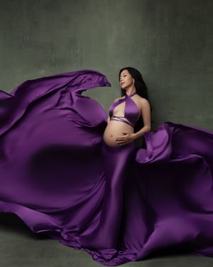 Stunning maternity portrait with flowing purple silk fabric against dark textured wall.