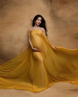 Glowing maternity photo with flowing mustard yellow dress against warm brown background.