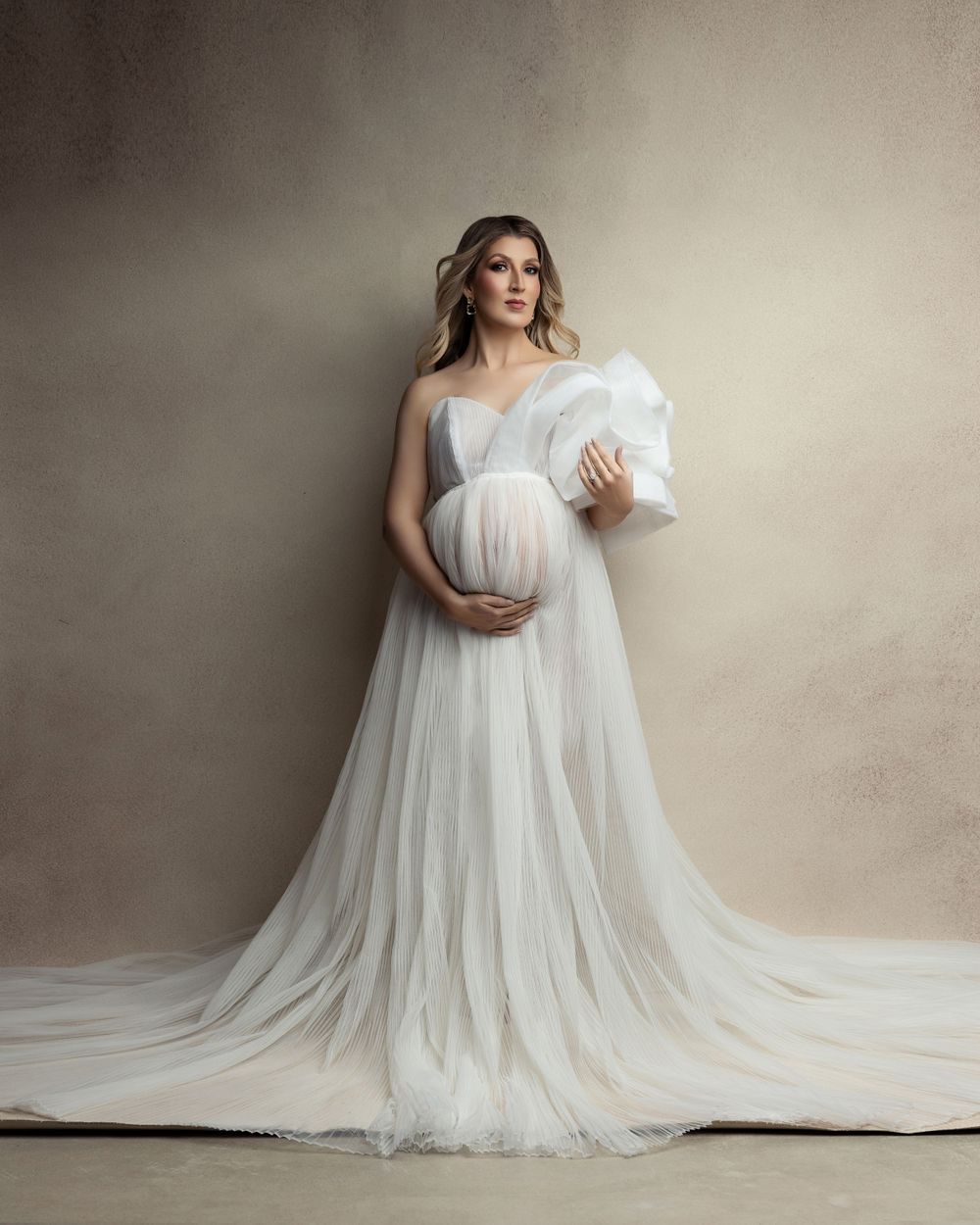 Elegant maternity portrait in flowing white empire waist gown against neutral backdrop.