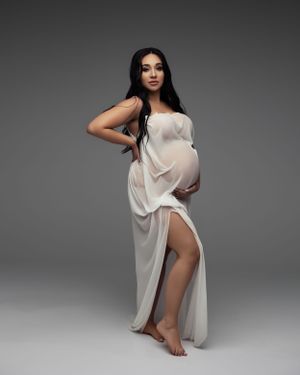 Studio maternity portrait in white flowing dress against grey backdrop.