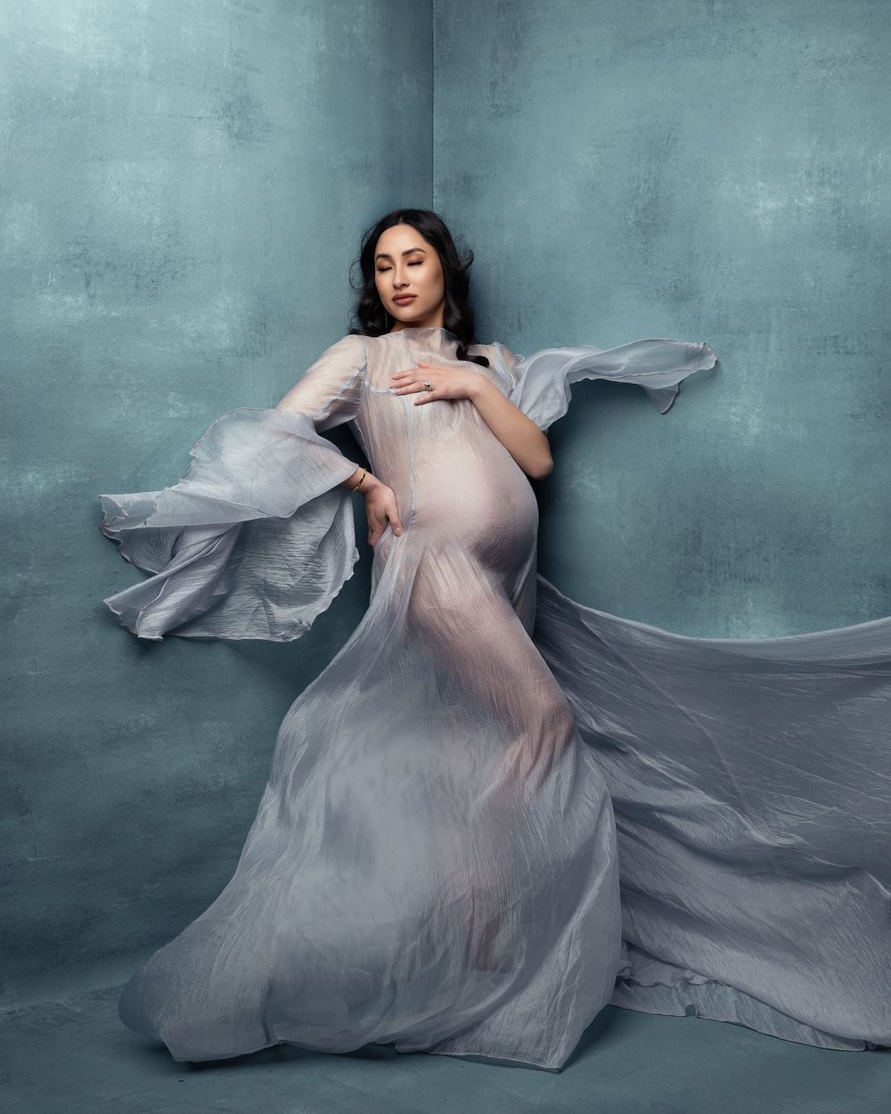Dramatic maternity portrait in flowing silver sheer gown against teal wall.