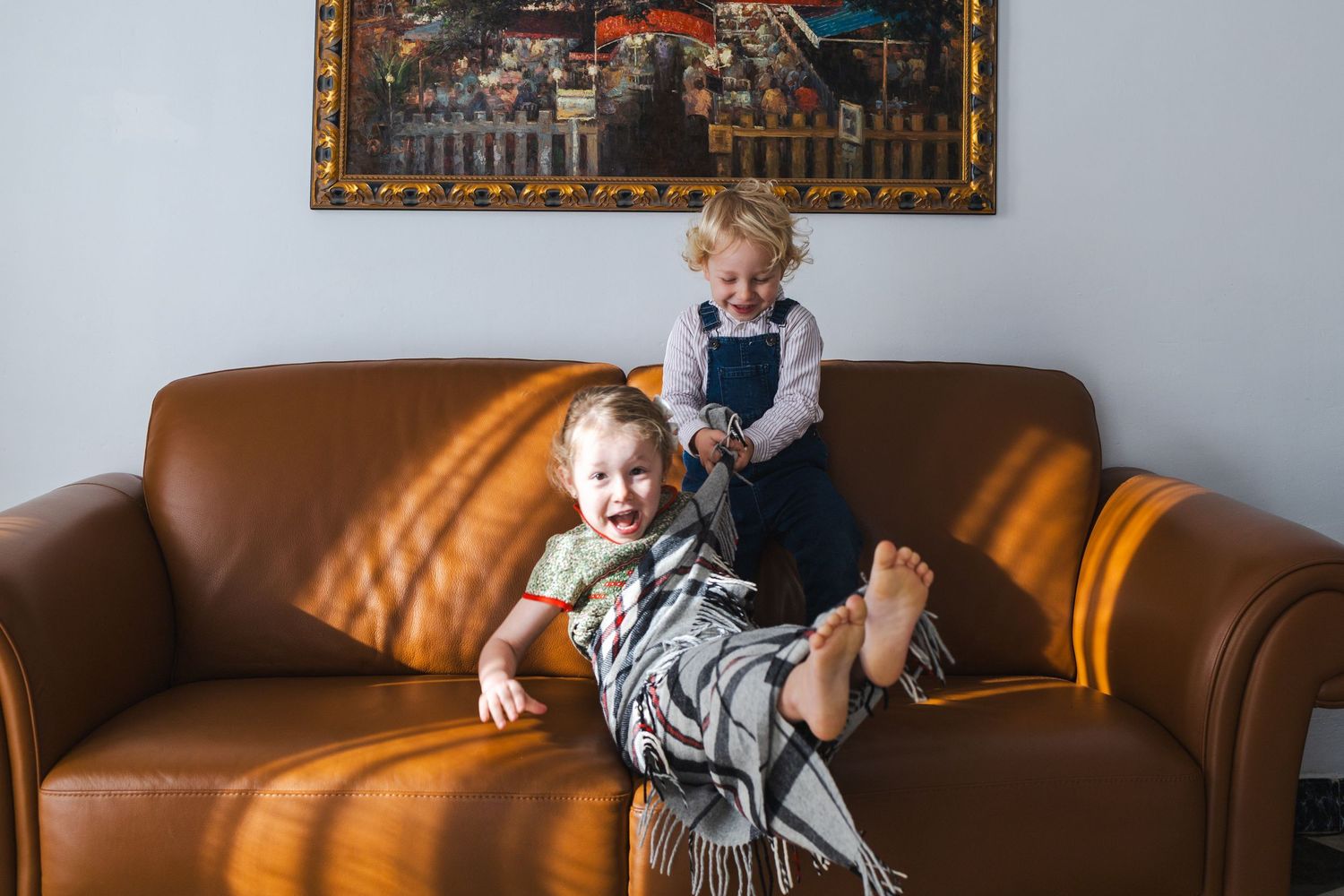 Two young children playing on a brown leather couch beneath a framed painting in a sunlit living room.