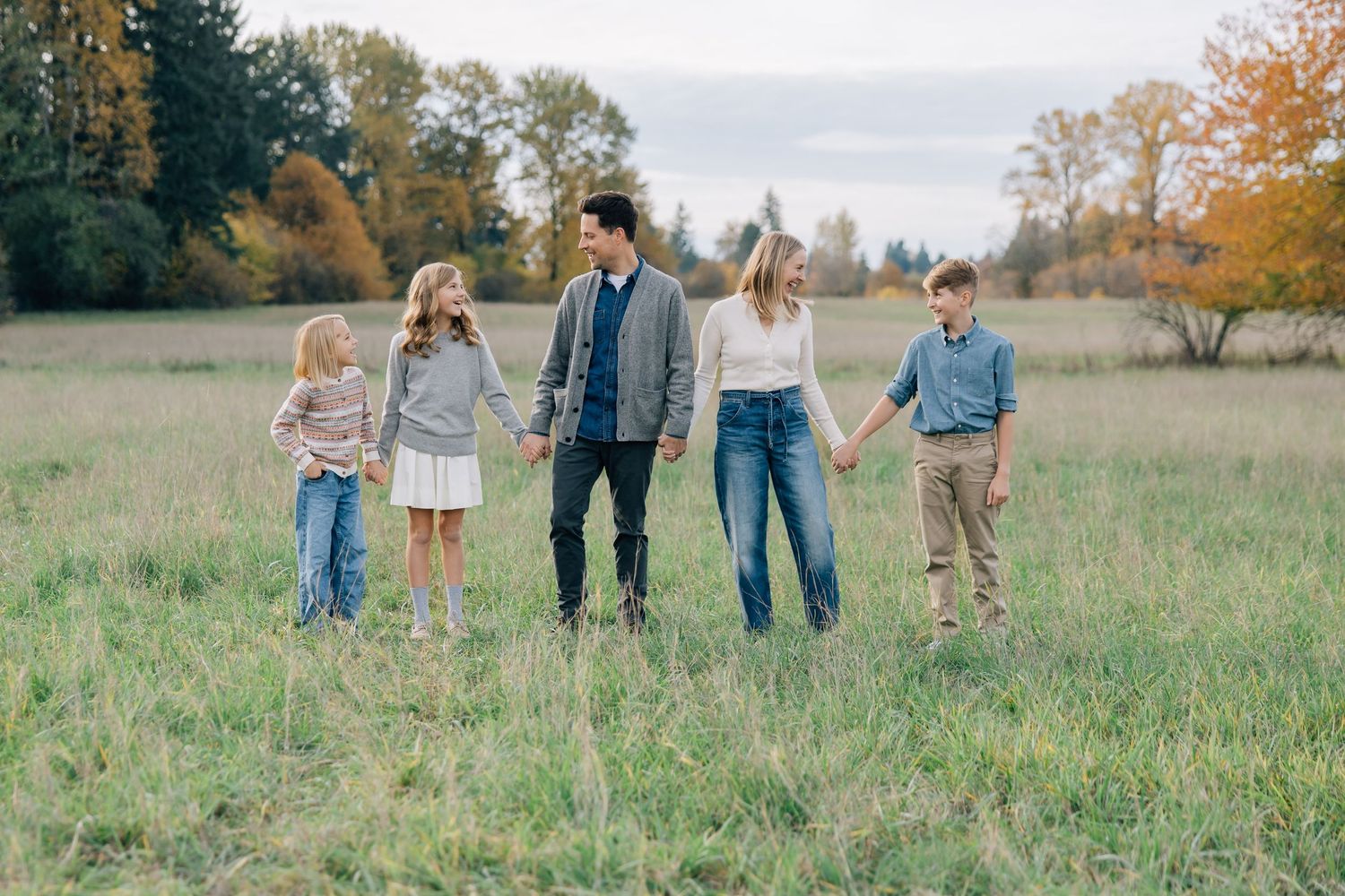 Family of five holding hands walking through a scenic autumn meadow with colorful trees in the background.