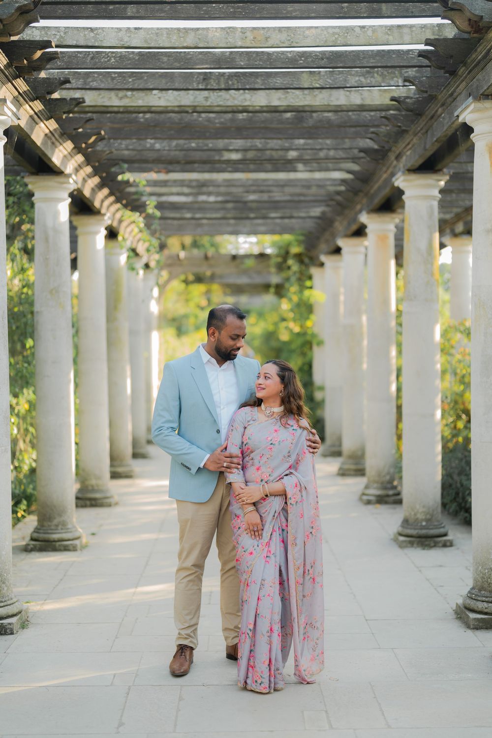 Couple in elegant attire walking under a stone pergola colonnade surrounded by lush greenery.