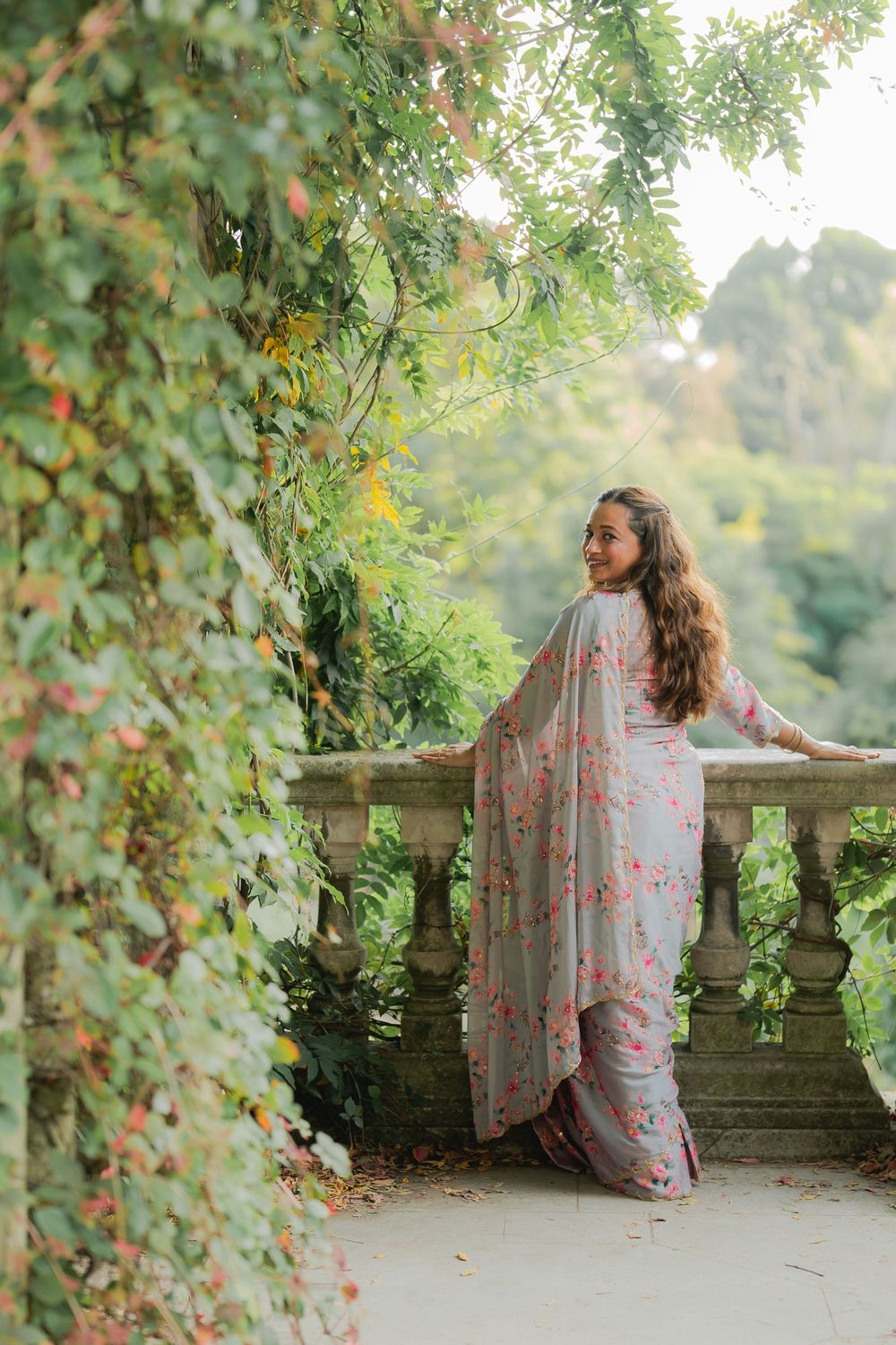 Woman in floral grey saree posing on stone balcony surrounded by lush green garden foliage.