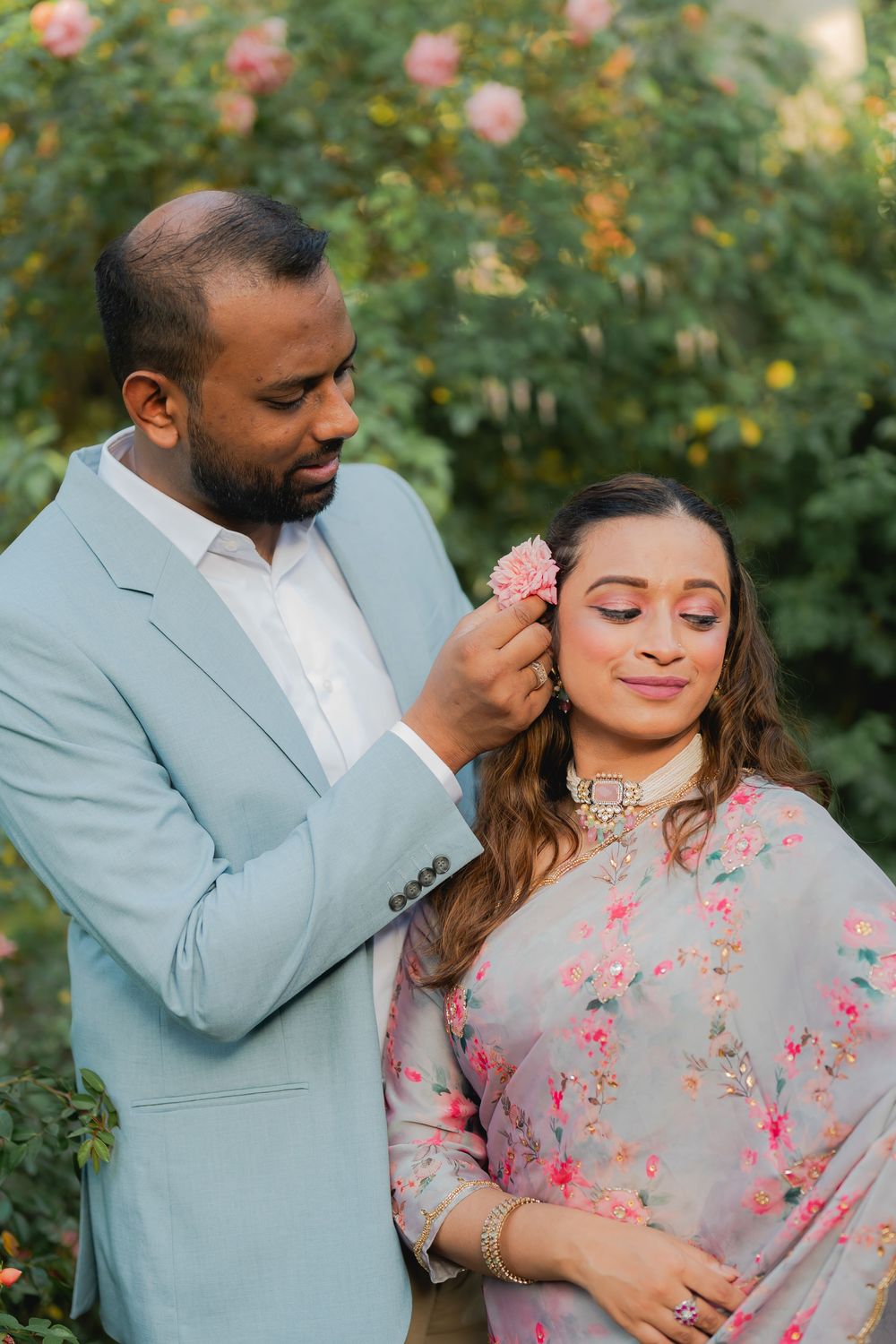 A man in a light blue suit places a pink flower in a woman's hair outdoors near blooming roses.