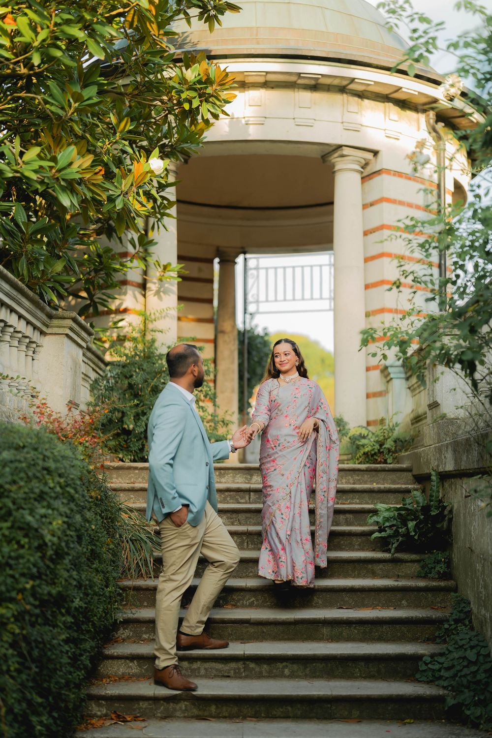 Couple in elegant attire on stone steps near classical columns, woman in pink saree, man in light blue blazer.