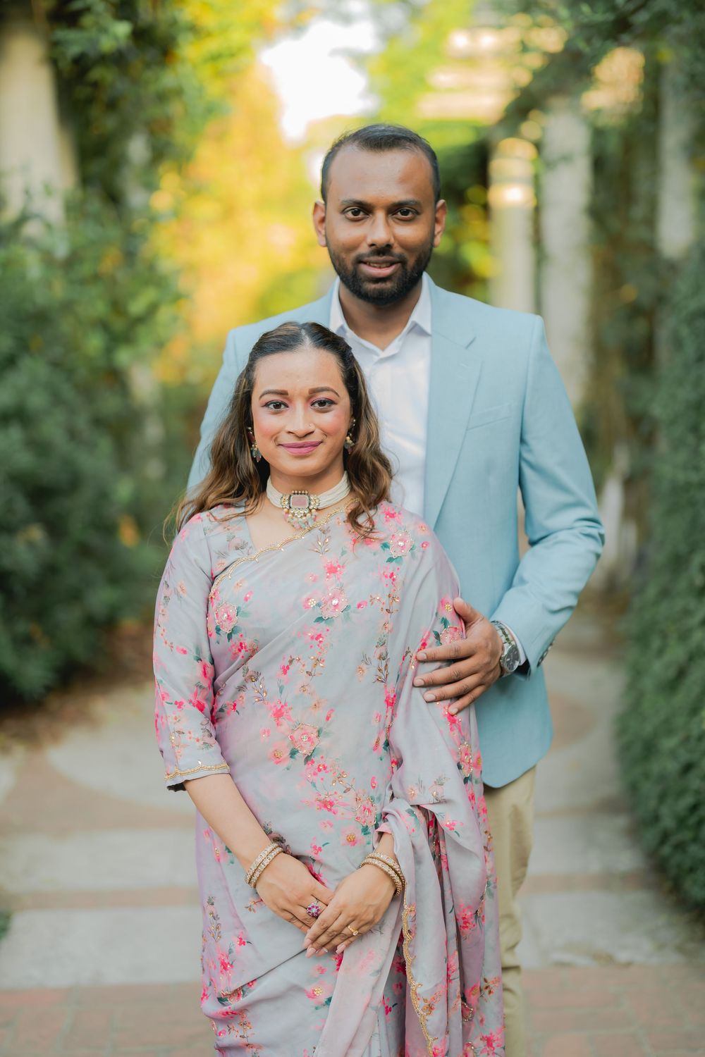 Couple in traditional Indian attire and light blue suit posing in a lush garden walkway.