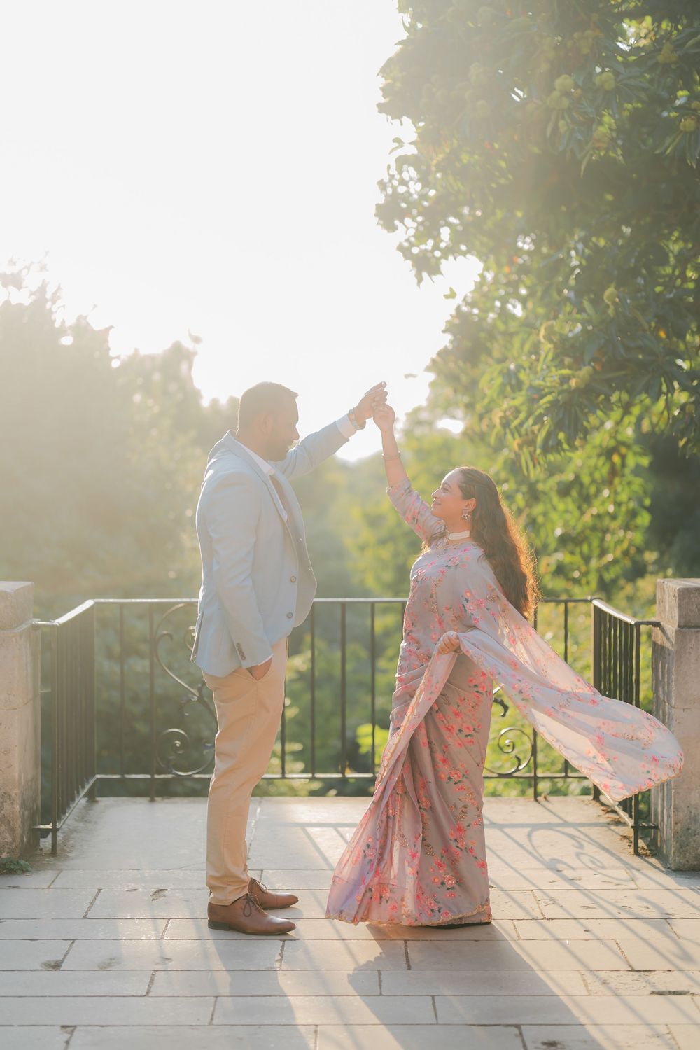 Couple dancing on a sunlit terrace, man in light blue suit twirling woman in flowing pink floral gown outdoors.