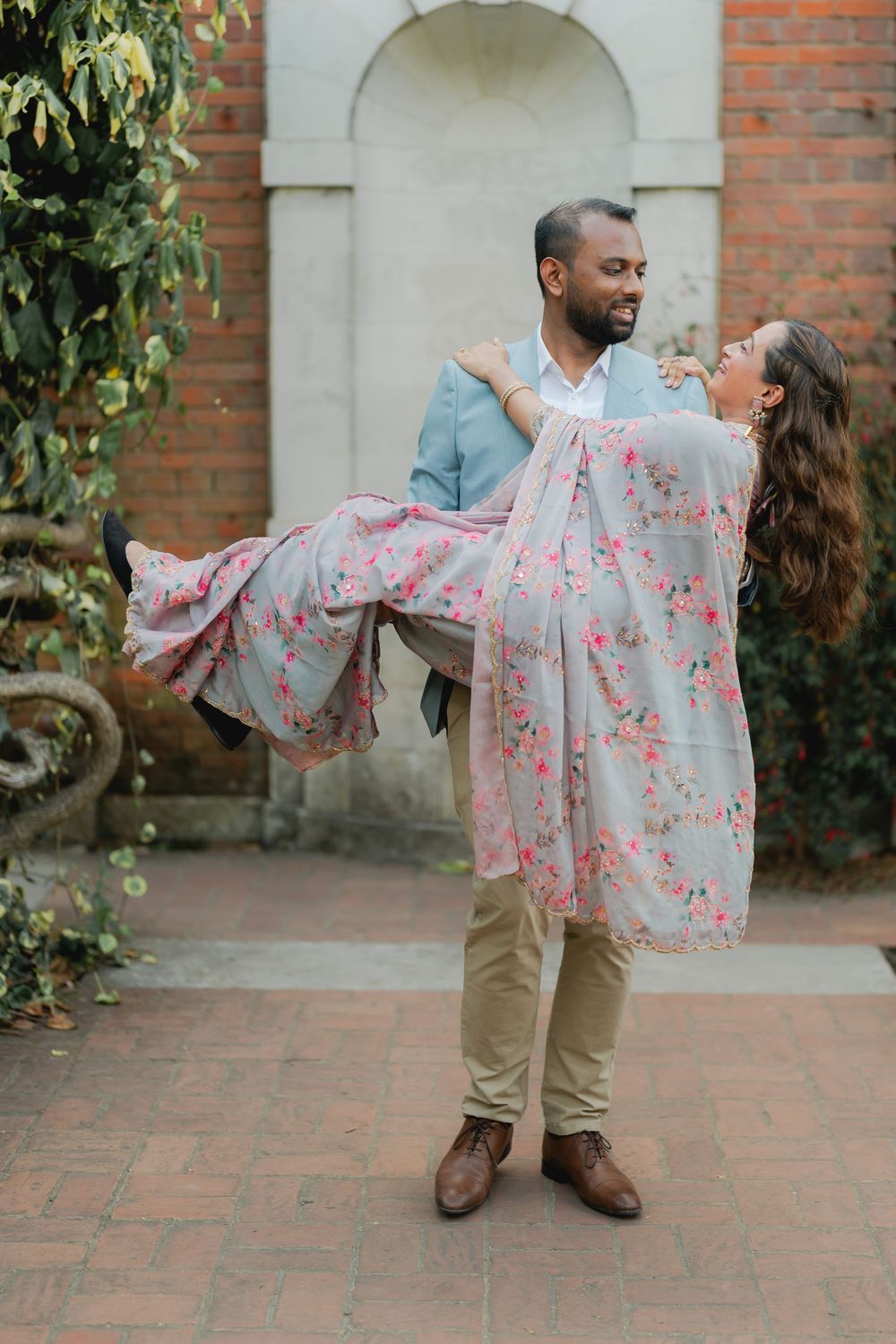 A man in a blue blazer lifts a woman in a floral gray dress in a romantic outdoor courtyard setting.