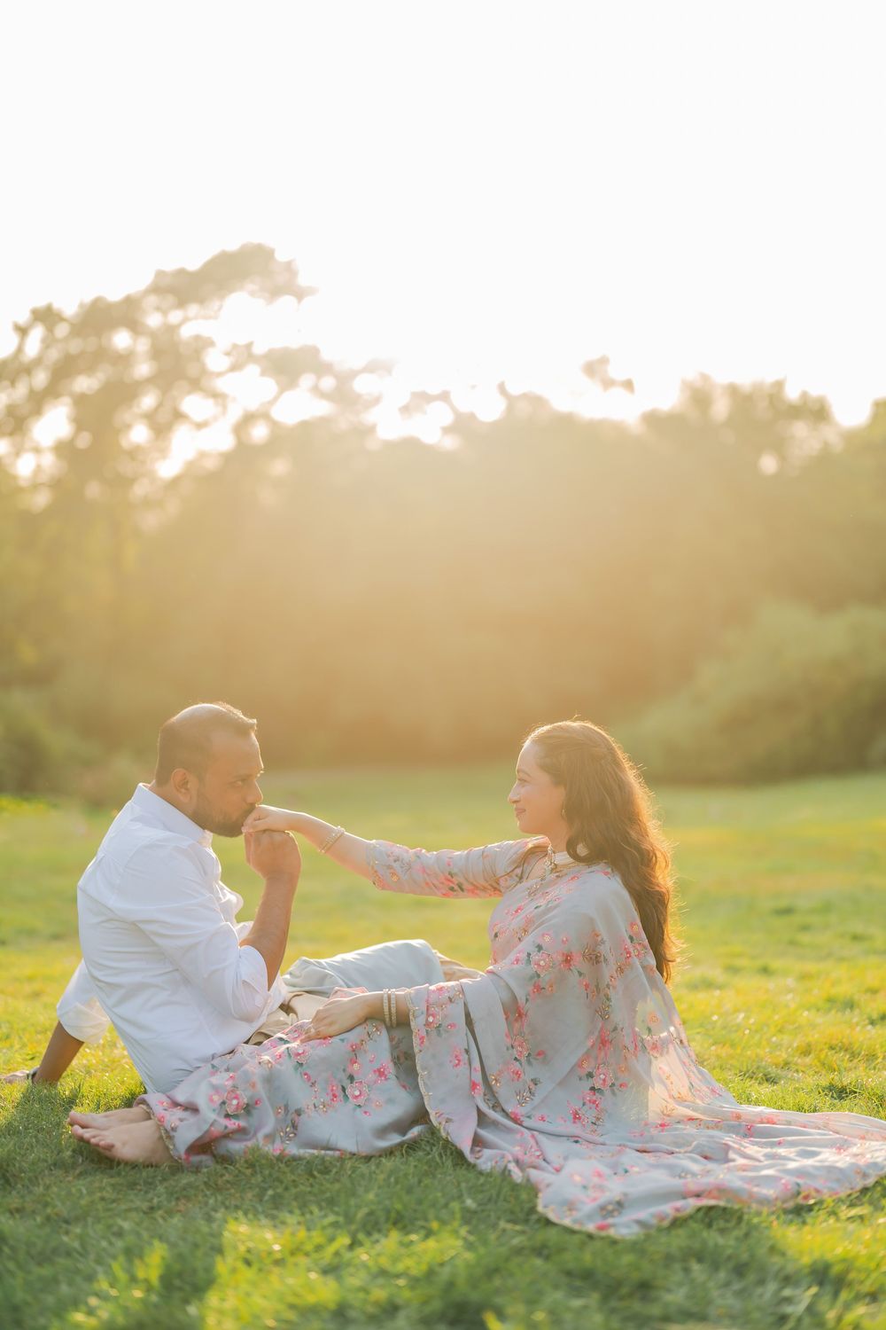 Couple sitting on grass at golden hour, man kissing woman's hand, both wearing floral outfits in a sunlit park.