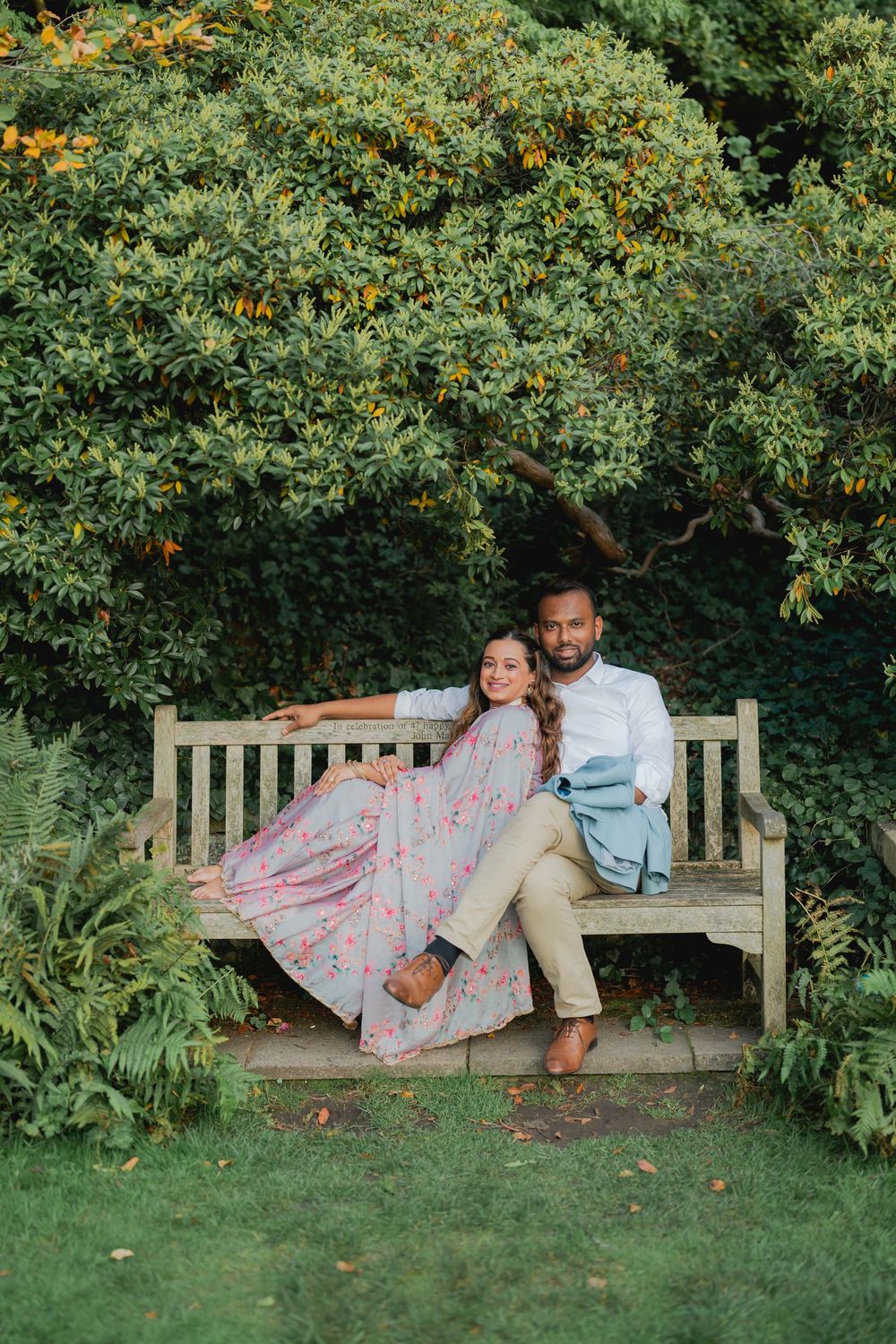 Couple sitting on a wooden garden bench surrounded by lush green shrubs, woman in floral dress leaning on man in white shirt.