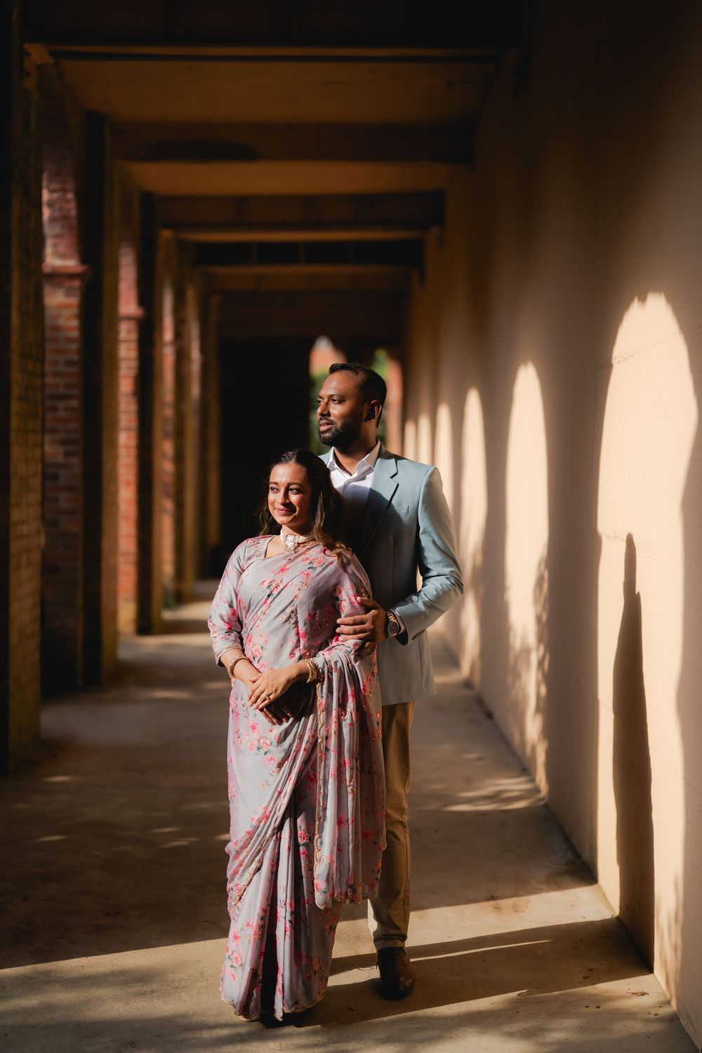Couple in traditional Indian attire walking through an elegant arched corridor with warm sunlight casting shadows.