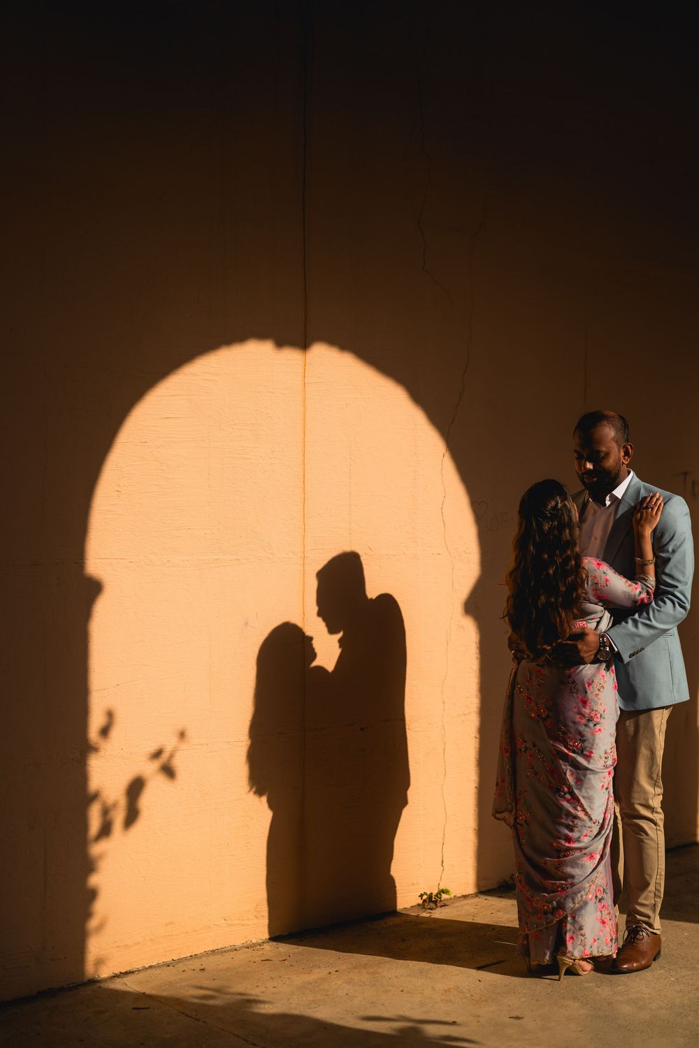 Couple casting romantic arch shadow on warm wall, woman in floral saree embracing man in suit at golden hour.
