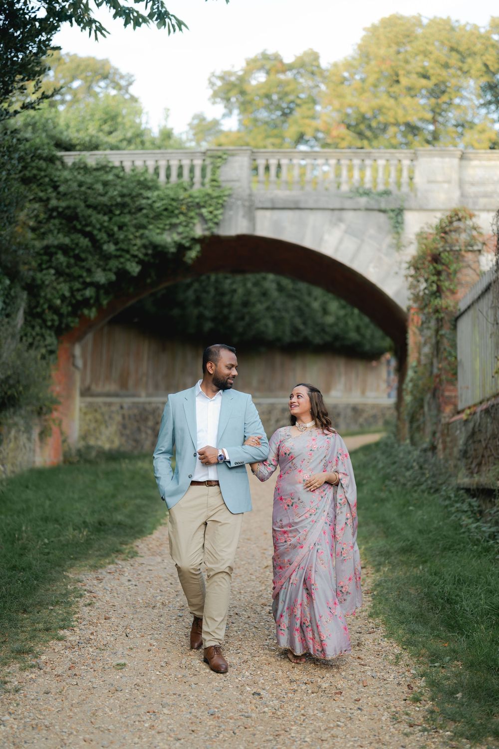 A couple walks hand-in-hand on a garden path, man in blue blazer, woman in pink floral saree, stone bridge behind.