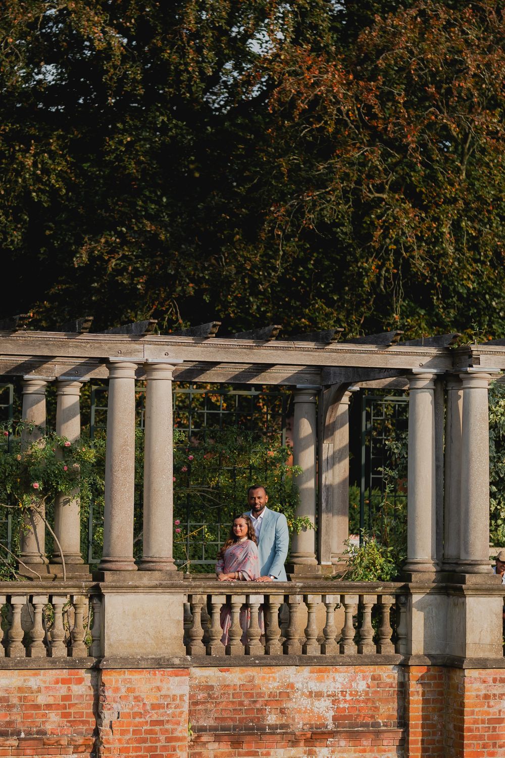 Couple standing under a stone pergola with classical columns above a brick balustrade, surrounded by lush green trees.