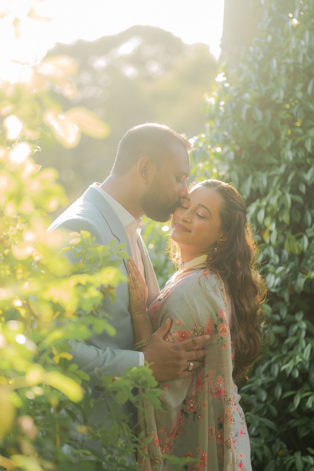 Couple sharing a romantic moment in a sunlit garden, man kissing woman's forehead in elegant traditional attire.