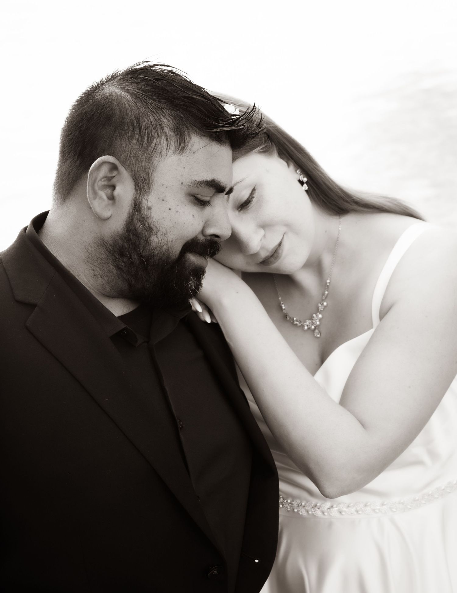 Black and white wedding photo of a bearded groom and bride in white dress tenderly touching foreheads in romantic pose.