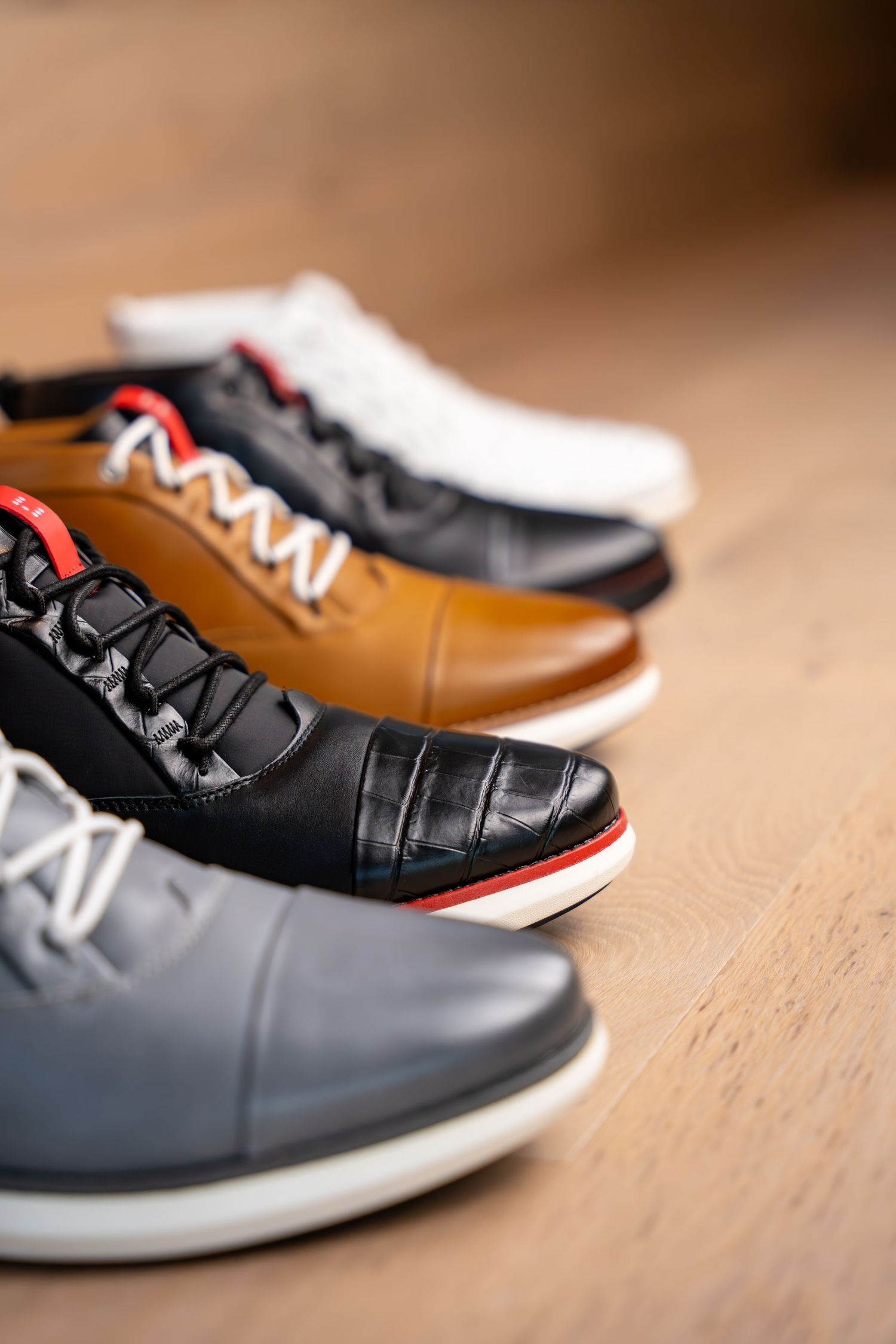 Four stylish leather sneakers in gray, black, tan, and white displayed in a row on a wooden floor.