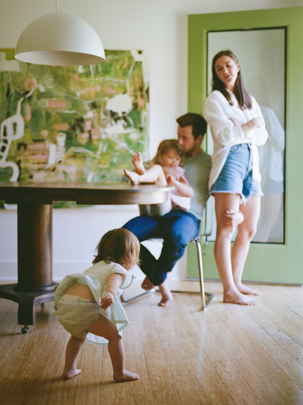 Family enjoying time at home, parents watching toddler play on hardwood floors near dining table.