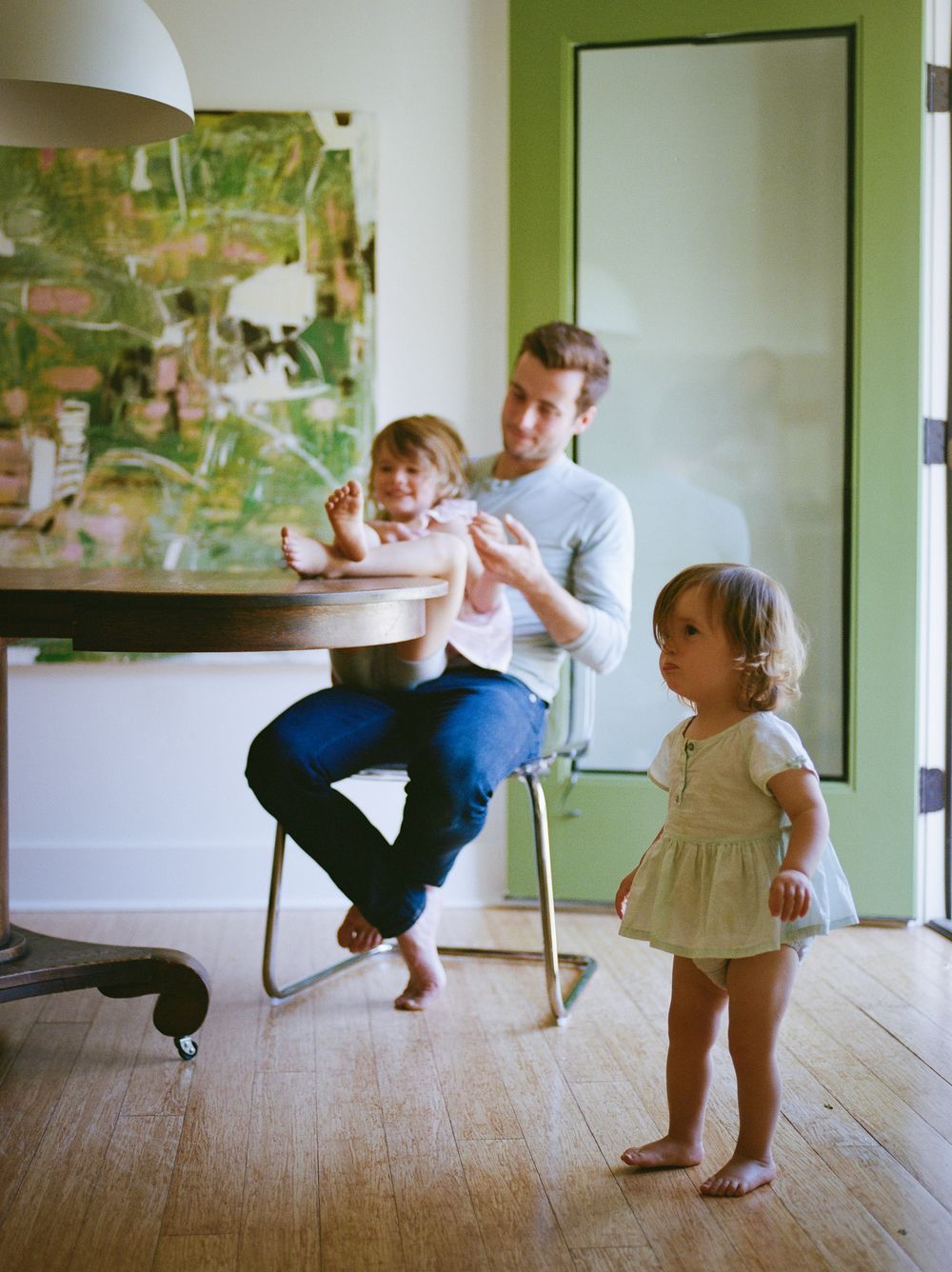 A father sits on a chair interacting with a toddler while another young child stands on hardwood floors indoors.