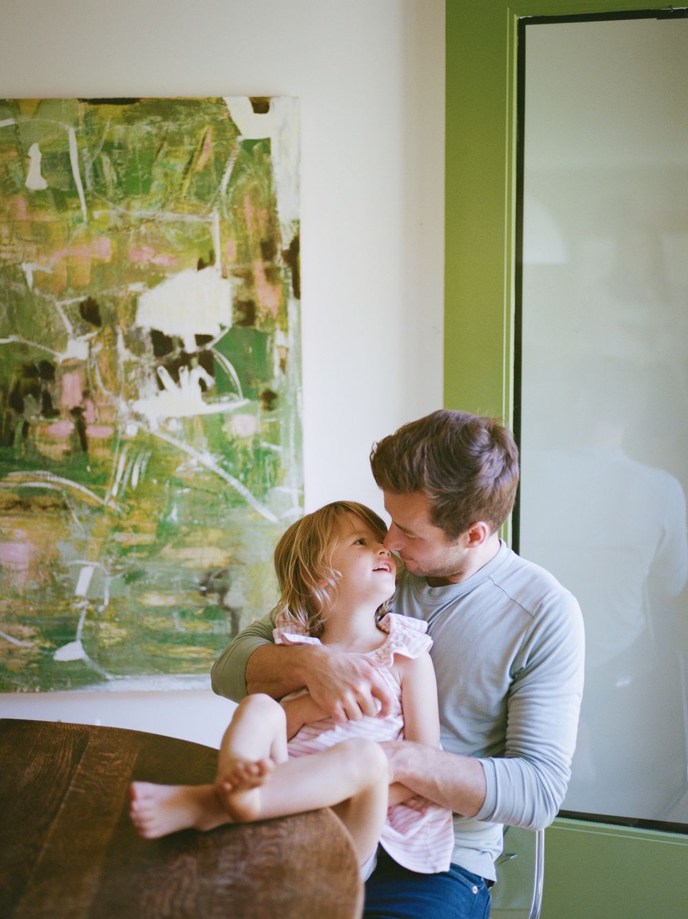 A father tenderly holds his laughing toddler daughter while sharing a sweet moment indoors near abstract wall art.