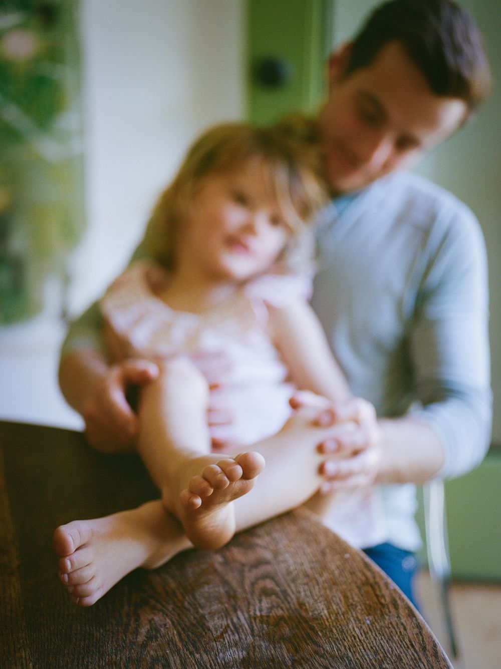 Father tenderly holds young toddler girl on wooden table, child's bare feet in focus in warm indoor setting.