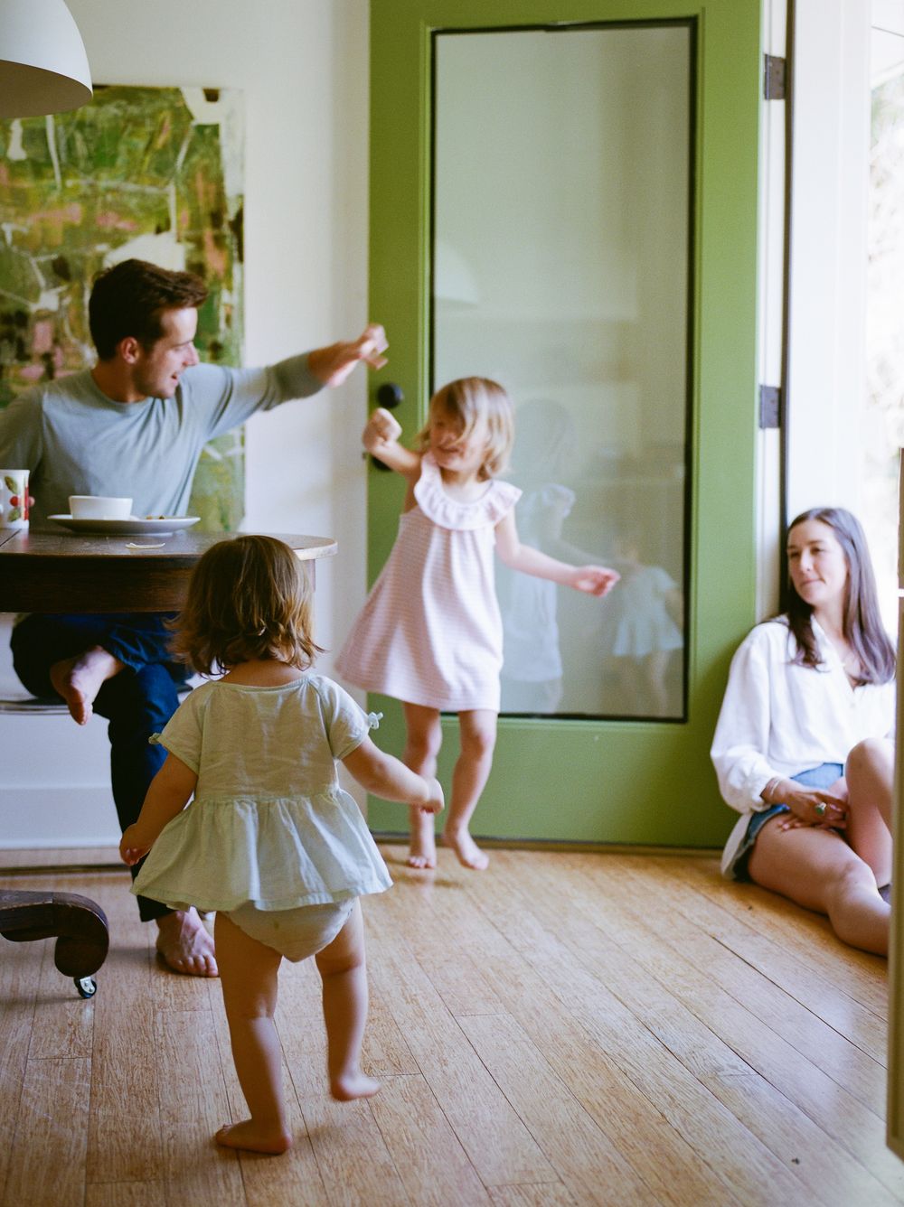 A family plays together indoors on hardwood floors as dad dances with two young girls while mom watches smiling.