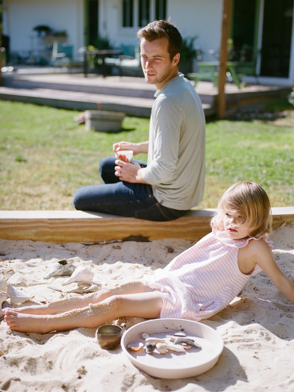 A man and young girl play together in a backyard sandbox on a sunny day.