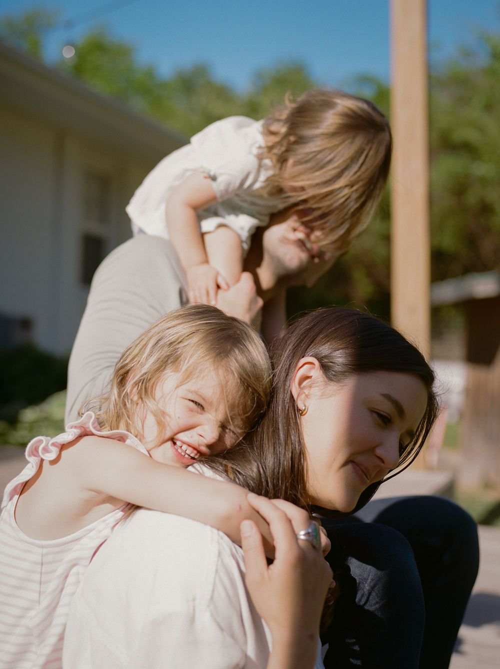 A mother smiles warmly as two young children climb and hug her outdoors.