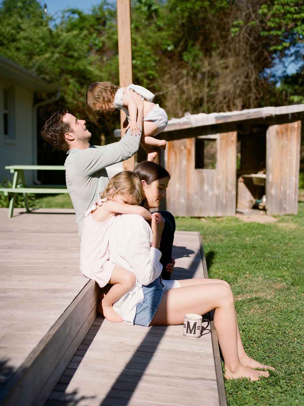 Father lifts toddler overhead while mother and child sit on outdoor wooden bench in sunny backyard.