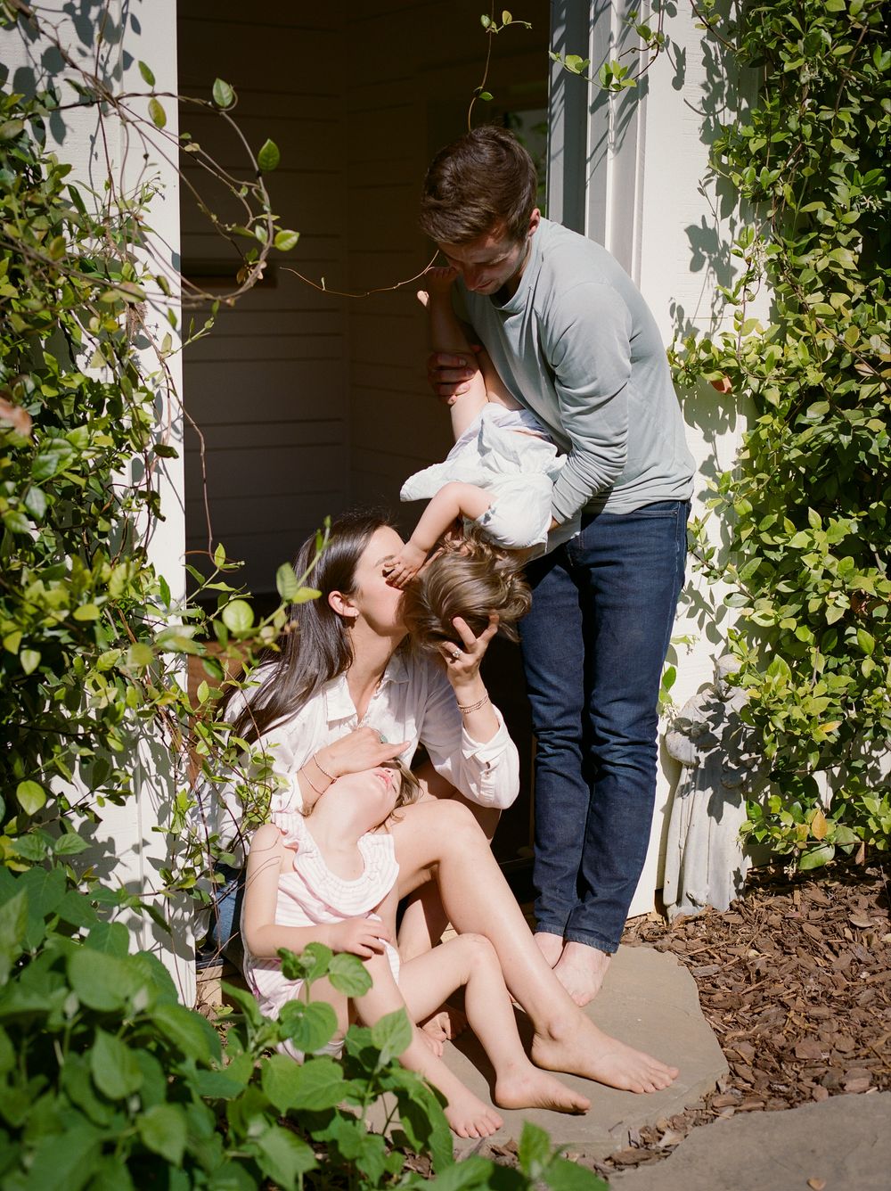A happy family of four shares a joyful moment together on a sunny home doorstep surrounded by lush greenery.