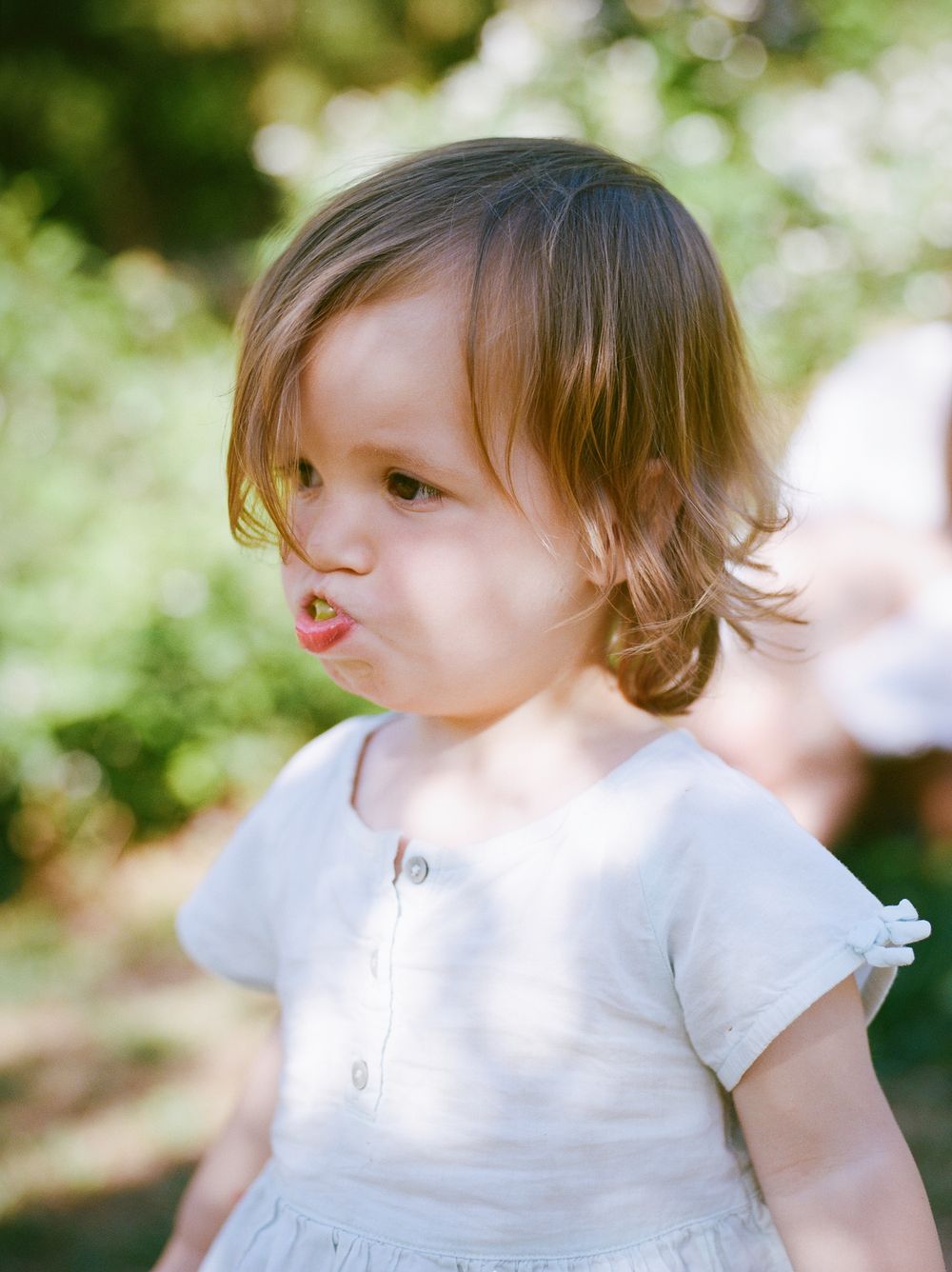 Adorable toddler girl with short brown hair wearing white dress makes a pouty face outdoors.