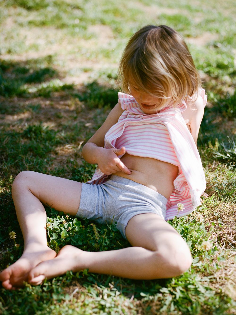 Young girl sitting barefoot on grass in a pink striped top and grey shorts, looking down on a sunny day.
