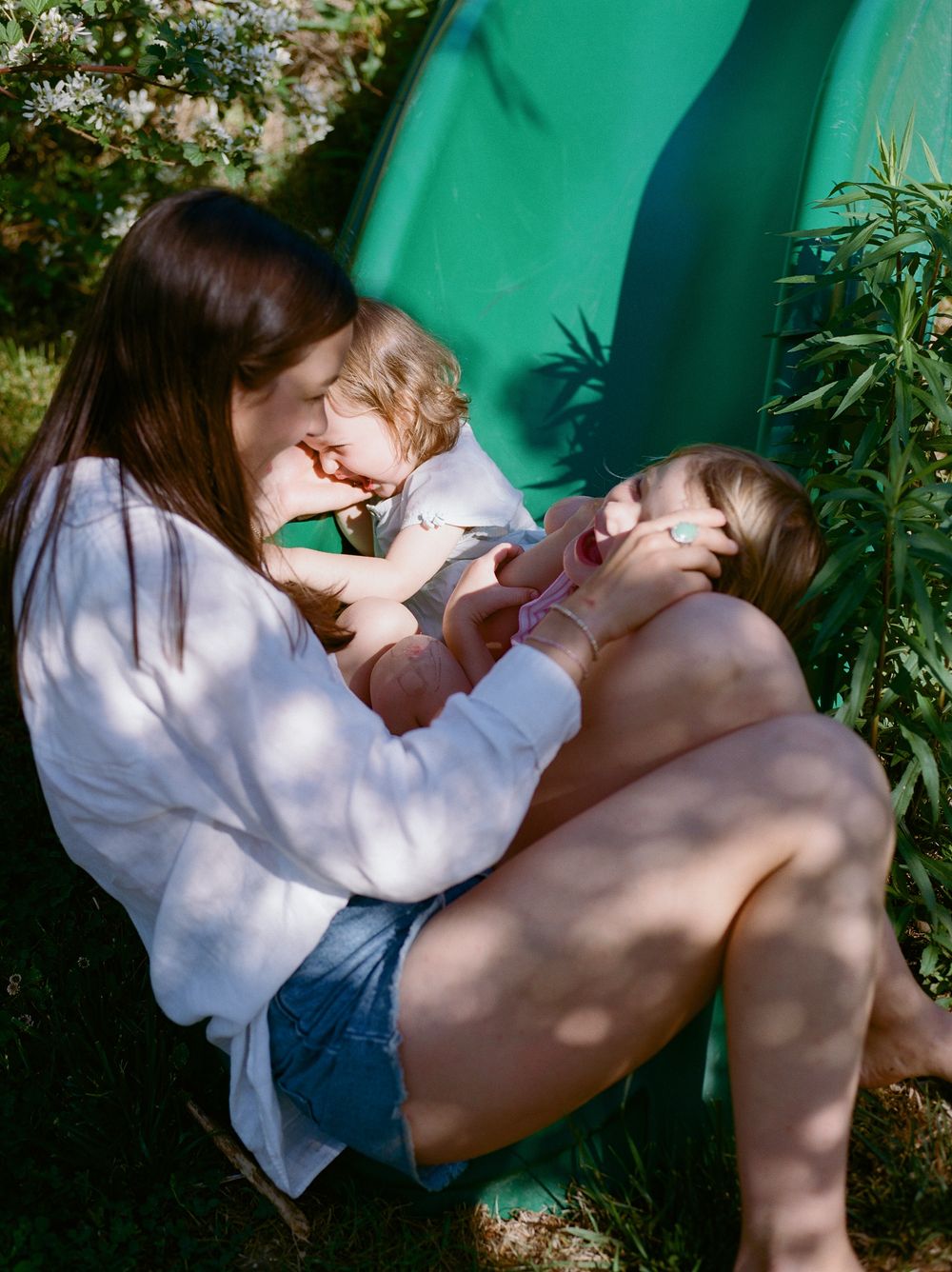 Mother sits outdoors cradling two young children in her lap near a green slide in garden.