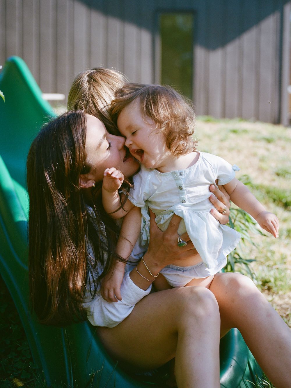 Joyful mother kissing laughing baby girl on green slide while older child watches from behind.