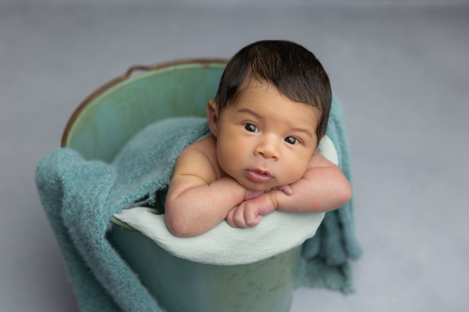 Newborn baby resting chin on hands inside a teal bucket with soft blanket, studio photography.