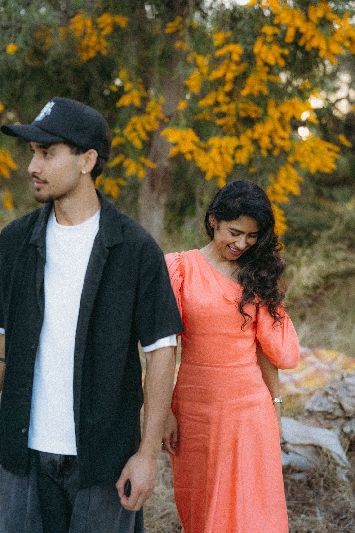 A young couple stands outdoors near yellow-bloomed trees, man in black shirt and cap, woman in coral dress smiling.