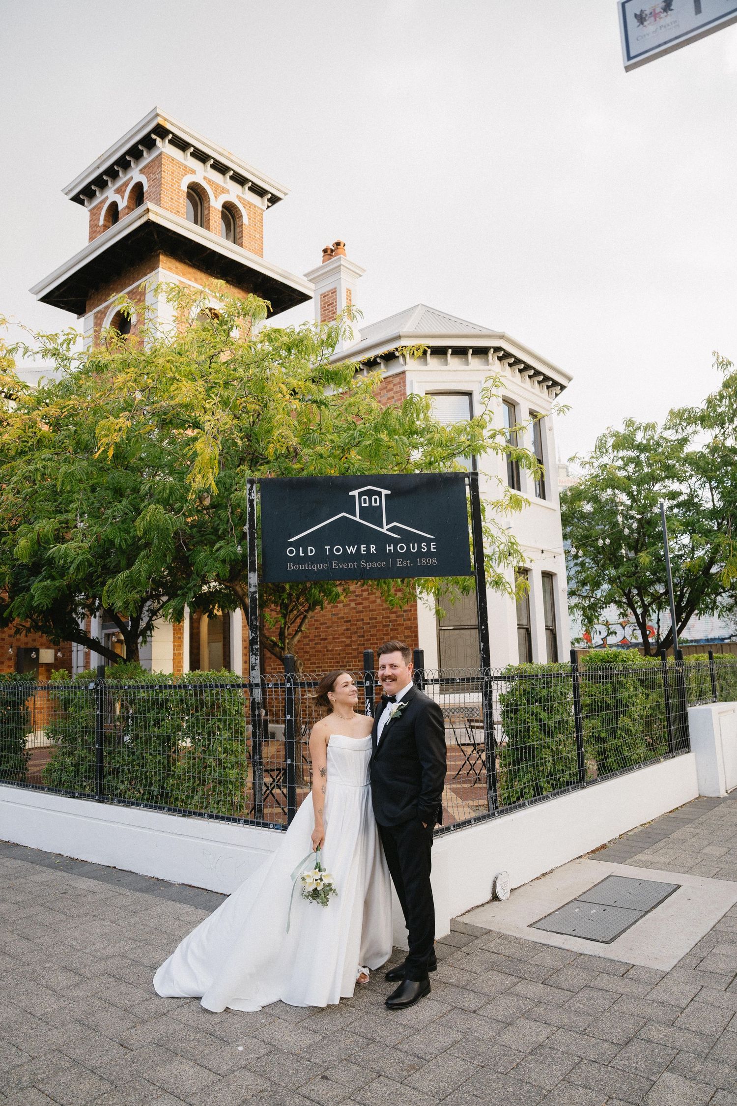 Bride and groom posing in front of Old Tower House venue, bride in white gown holding bouquet, groom in black tuxedo.